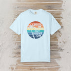A light blue short-sleeve t-shirt with a circular retro graphic on the chest, laid flat on a rustic wooden plank background. The graphic features a sunset gradient in orange, yellow, green, and blue with a palm tree silhouette and the text 'BREATHE IN BREATHE OUT MOVE ON'.