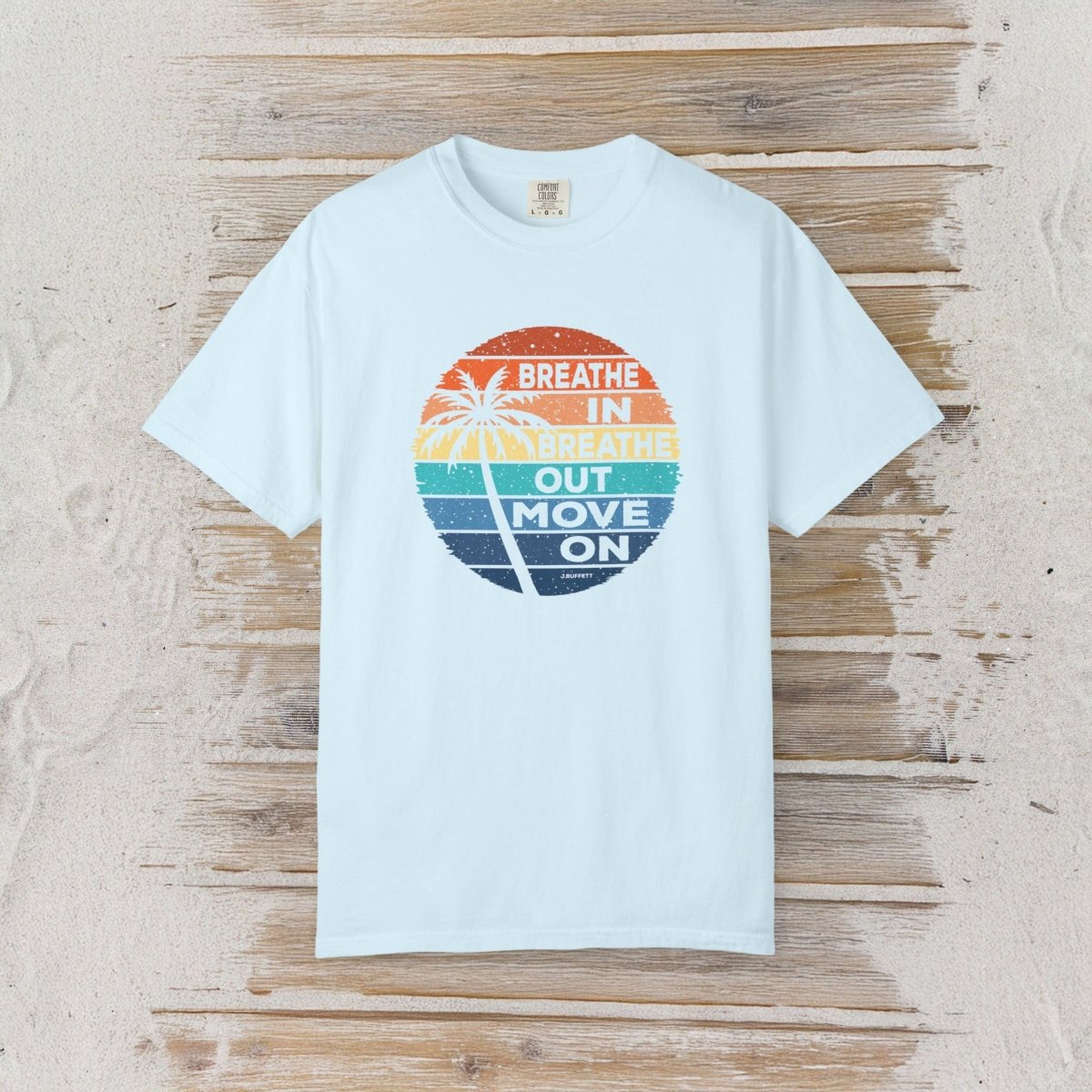 A light blue short-sleeve t-shirt with a circular retro graphic on the chest, laid flat on a rustic wooden plank background. The graphic features a sunset gradient in orange, yellow, green, and blue with a palm tree silhouette and the text 'BREATHE IN BREATHE OUT MOVE ON'.