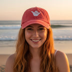 A smiling woman with long wavy red hair wearing a coral pink baseball cap with a white hand gesture logo, standing on a beach at sunset with the ocean in the background.