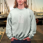 Person wearing a light gray "It's 5 O'Clock Somewhere" Sweatshirt with text standing on a dock with boats in the background