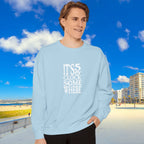 Person wearing a light blue sweatshirt with text, standing in front of a beach and buildings.