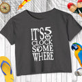 Black t-shirt with 'It's 5 o'clock somewhere' text on a wooden surface with decorative items.