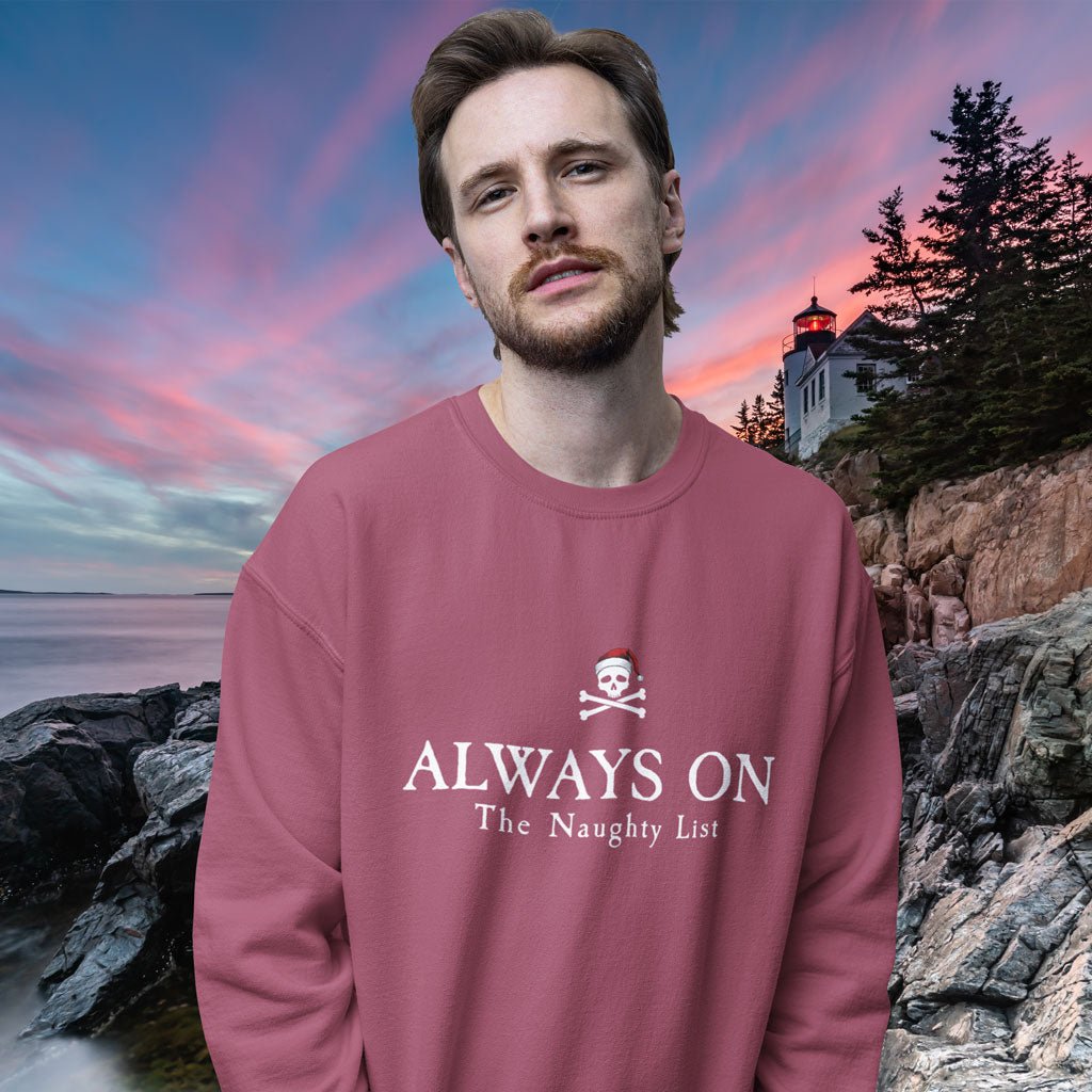 Man wearing a pink sweatshirt with 'ALWAYS ON The Naughty List' text and skull design, standing against a scenic background with a lighthouse.
