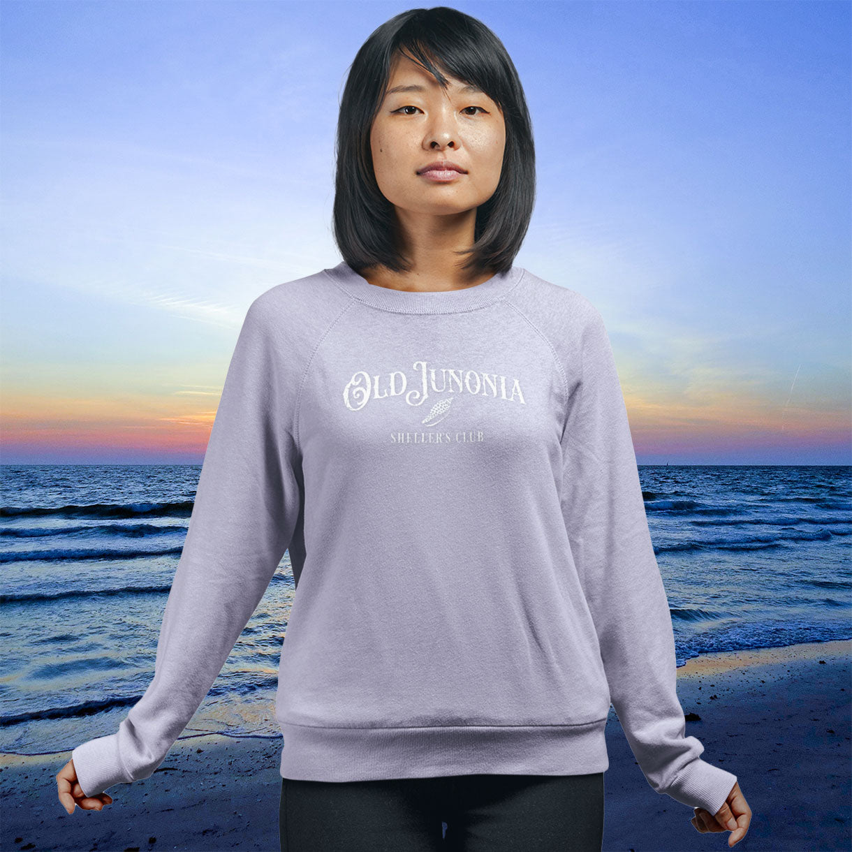 Person wearing a gray sweatshirt with 'Old Junonia' logo on a beach at sunset