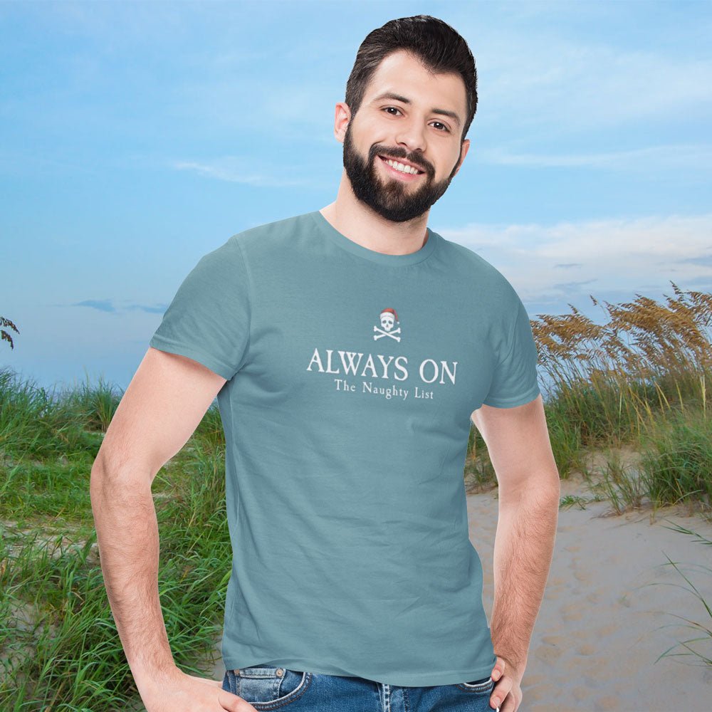 Man wearing a teal t-shirt with 'ALWAYS ON The Naughty List' text on a beach.