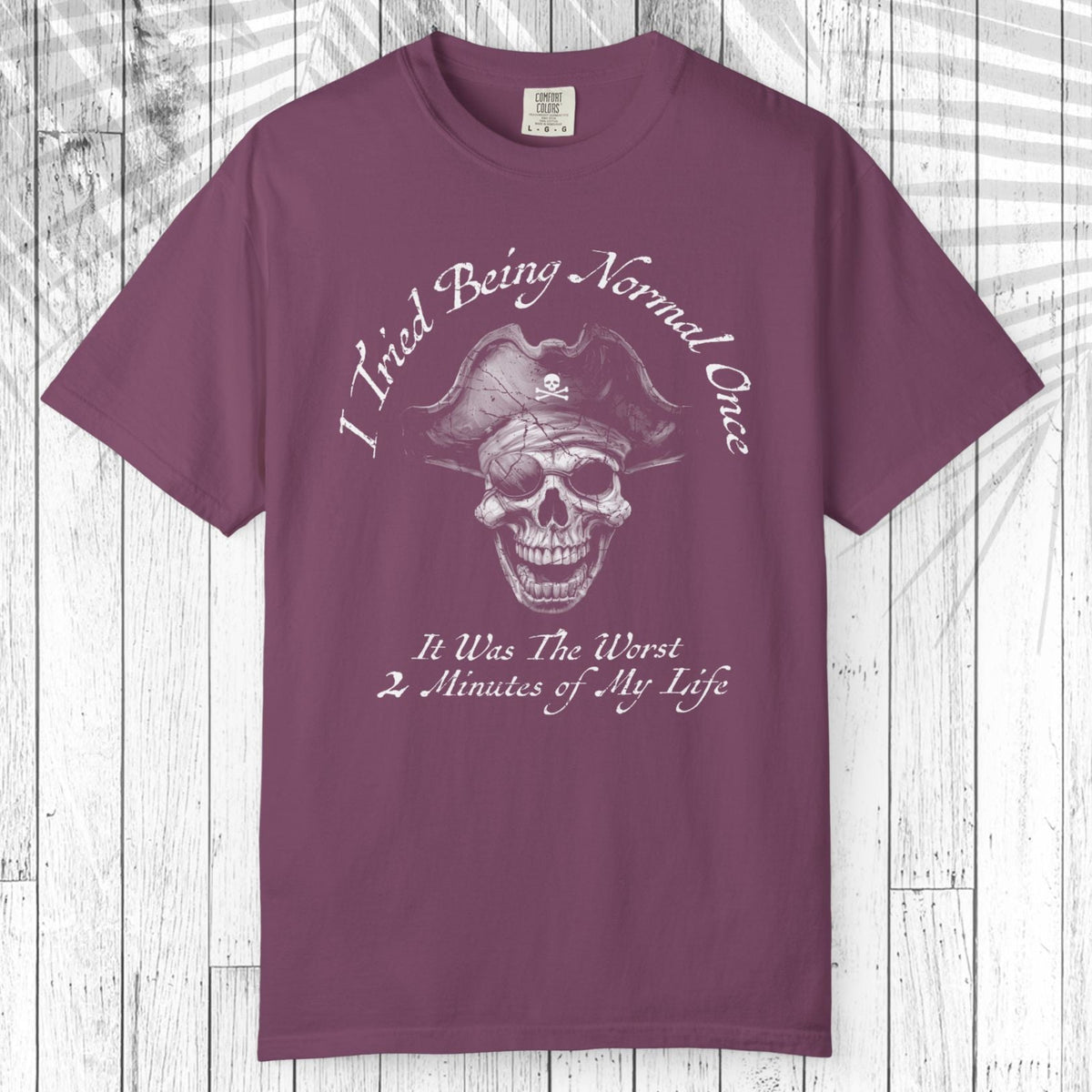A short-sleeve crewneck t-shirt in a dusty plum purple color, featuring a white graphic of a skull wearing a pirate hat with the text 'I Tried Being Normal Once' above and 'It Was The Worst 2 Minutes of My Life' below. The t-shirt is displayed flat against a white wood plank background.
