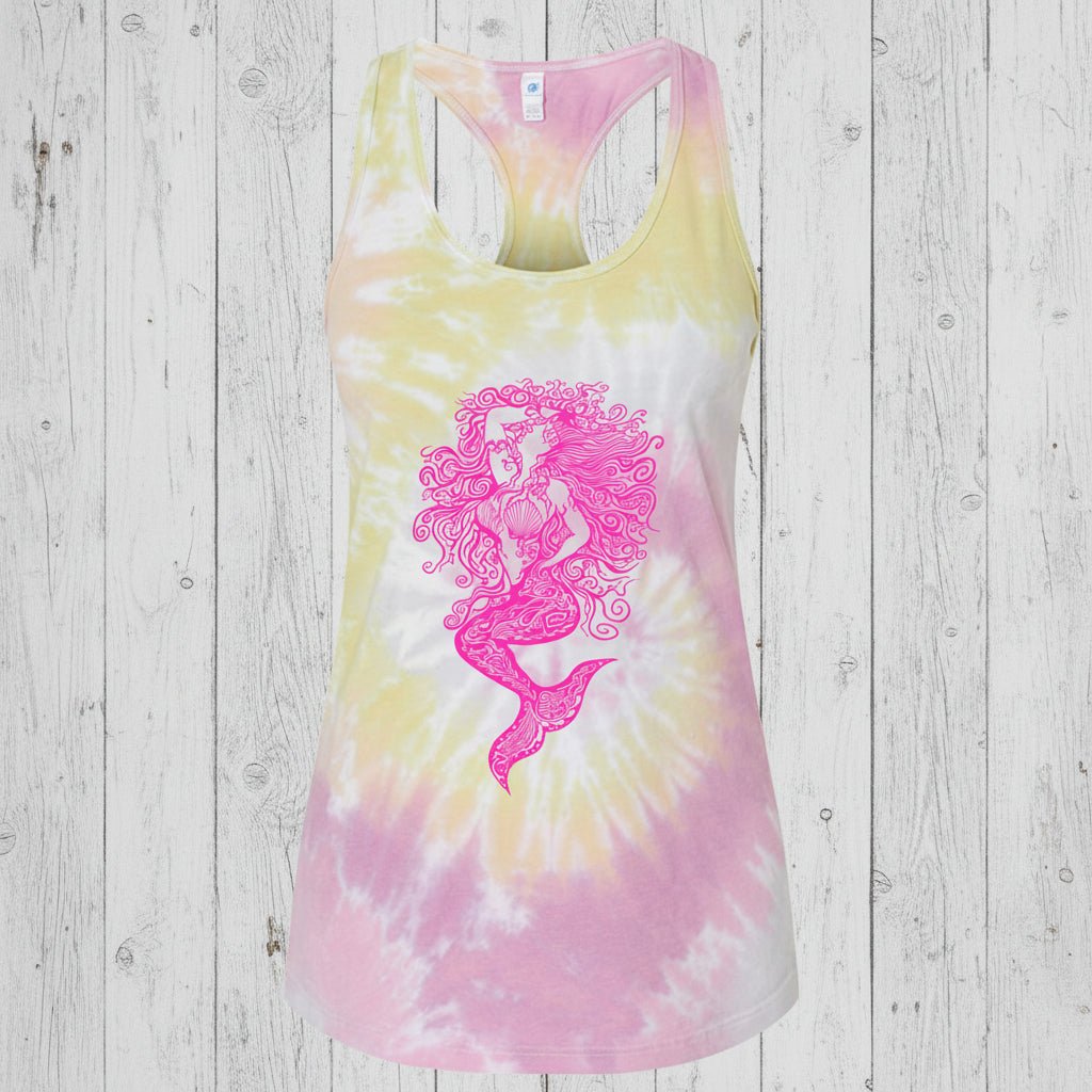 A women's racerback tank top featuring a pink mermaid graphic on a pastel tie-dye background of yellow, pink, and white, shown against a plain rustic wood background.