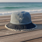 A bucket hat with a light blue denim-like crown, a dark navy brim with frayed edges, and a small white palm tree embroidery, resting on a wooden boardwalk with the ocean in the background.