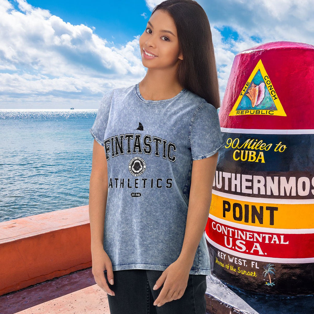 Woman wearing a 'Fintastic Athletics' t-shirt by the ocean with a colorful sign in the background.