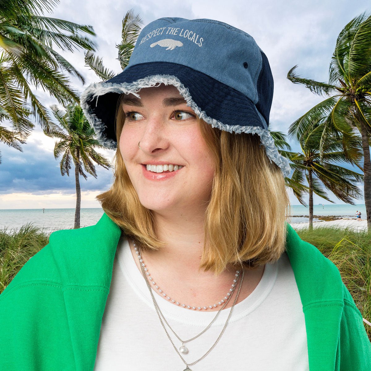 Person wearing a blue Embroidered Manatee bucket hat with text, standing in front of palm trees and the ocean.