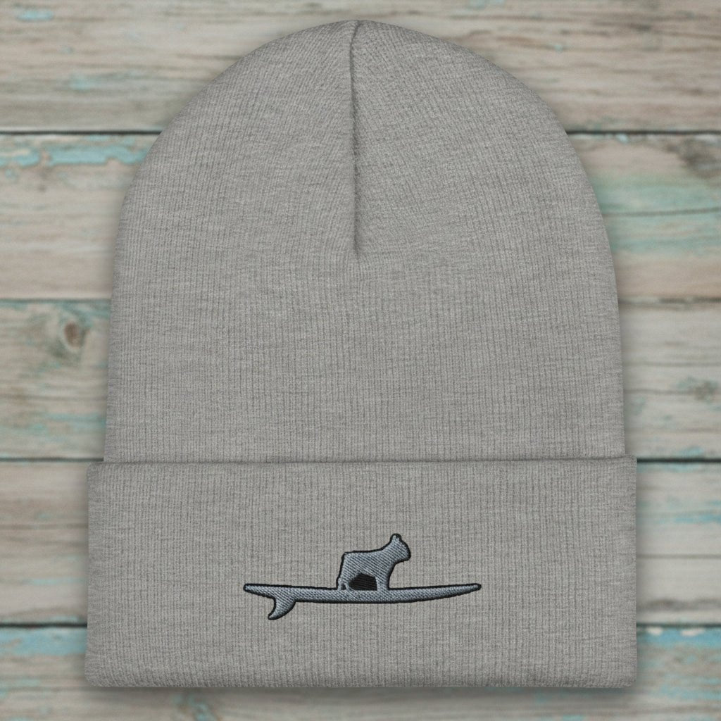 A light gray ribbed knit beanie hat with a folded cuff, featuring a small embroidered graphic of a frenchie standing on a surfboard on the front cuff.