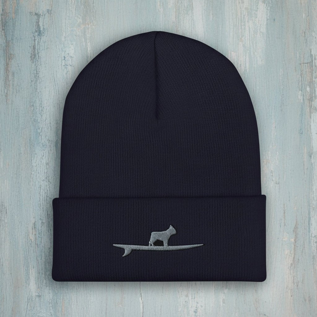 A dark navy blue ribbed knit beanie hat with a folded cuff, featuring a small gray embroidered graphic of a Frenchton Bulldog standing on a surfboard on the front.