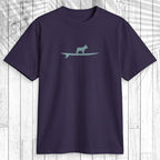 A dark purple short-sleeve t-shirt with a light teal graphic of a dog standing on a surfboard on the chest, displayed flat against a white wood plank background.