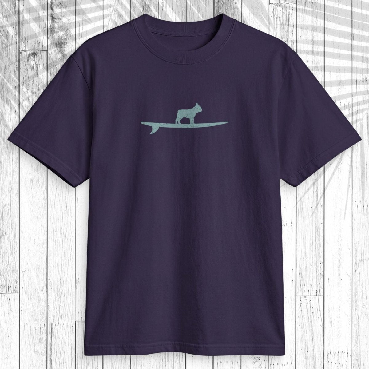 A dark purple short-sleeve t-shirt with a light teal graphic of a dog standing on a surfboard on the chest, displayed flat against a white wood plank background.