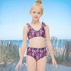 Young girl wearing a colorful seahorse-patterned two-piece swimsuit on a sand dune with a coastal style wood finc in the background