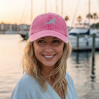 A smiling blonde woman wearing a pink baseball cap with a light gray shark logo, photographed outdoors at sunset near a marina with boats and water in the background.