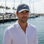 A man wearing a gray baseball cap with a white shark logo and a white button-down shirt, standing in front of a marina background with water and boats.