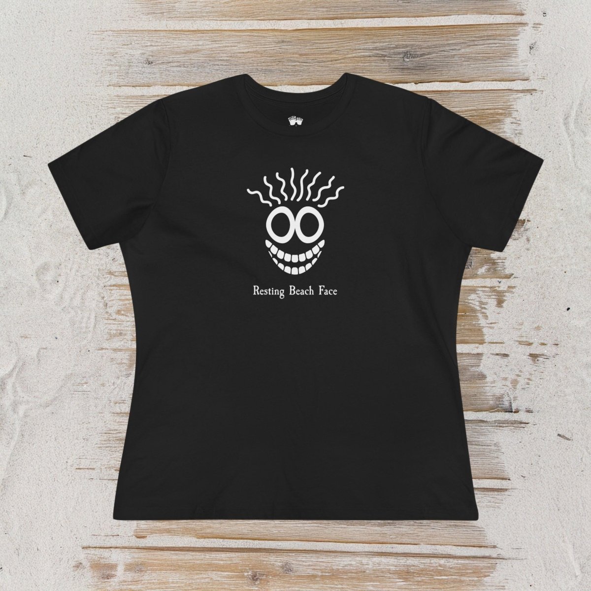 A black short-sleeve crewneck t-shirt laid flat on a light-colored, rustic wood plank background. The shirt features a white graphic of a skull-like face with wavy hair and the text 'Resting Beach Face' printed below it.