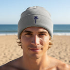 A heather gray knit beanie hat with a folded cuff and a small navy blue palm tree graphic on the front, shown young man on Havens Beach