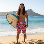 Pacific Islander dude with long hair wearing tropical palm print board shorts, holding a surfboard on a Hawaiian Island Beach. 