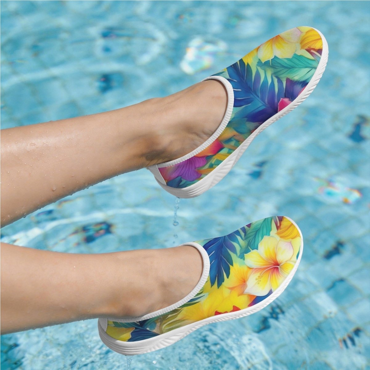 A pair of slip-on water shoes with a vibrant tropical floral and leaf print in yellow, blue, green, and purple, shown being worn with legs extended over a swimming pool.