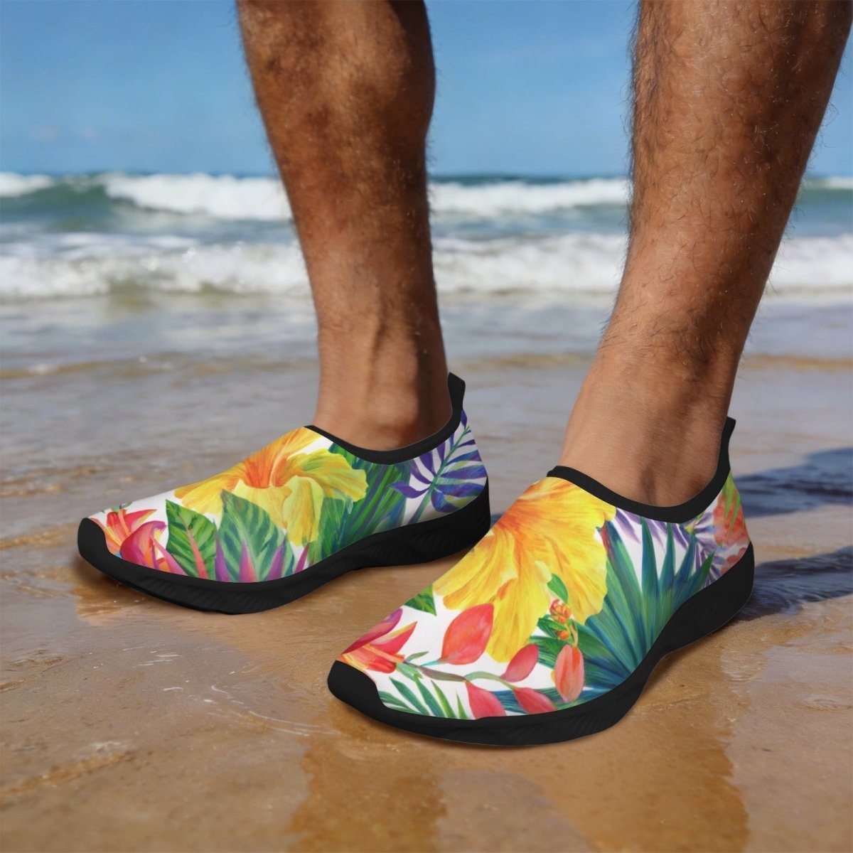 A person standing on a sandy beach wearing slip-on water shoes with a bright tropical floral and leaf print in yellow, green, pink, orange, and blue, with black soles. The ocean and blue sky are visible in the background.