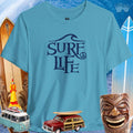 Surfer Life Tee in turquoise, featuring surf-themed design, surrounded by beach and surf accessories.