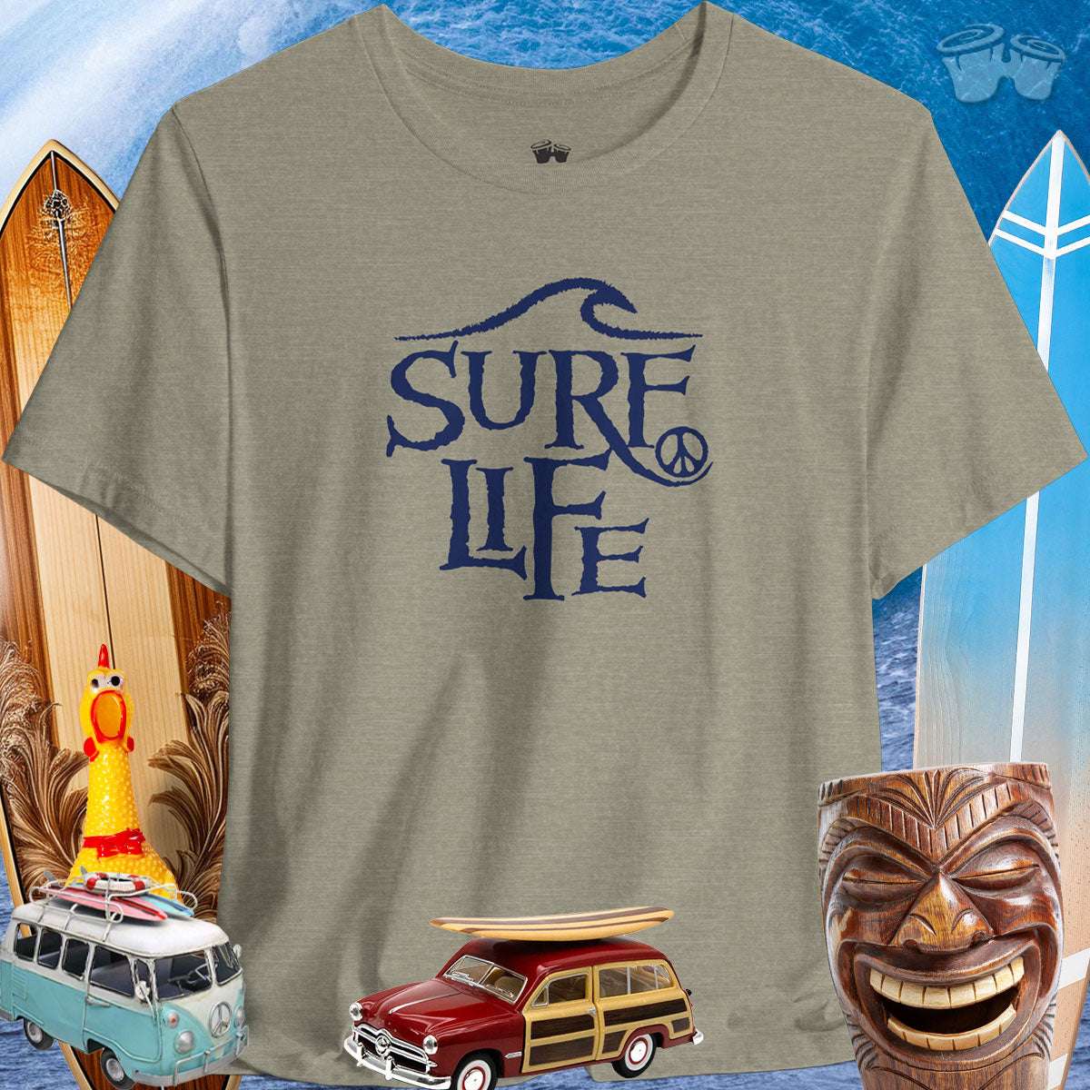 Surfer Life Tee in muted green with surf-themed graphics, surrounded by vintage cars and tiki decor, perfect for beach lovers.