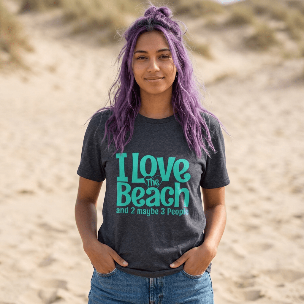 A woman with purple hair stands on a sandy beach wearing a dark heather gray Clever T-Shirt t-shirt with a teal graphic that reads 'I Love The Beach and 2 maybe 3 People'.