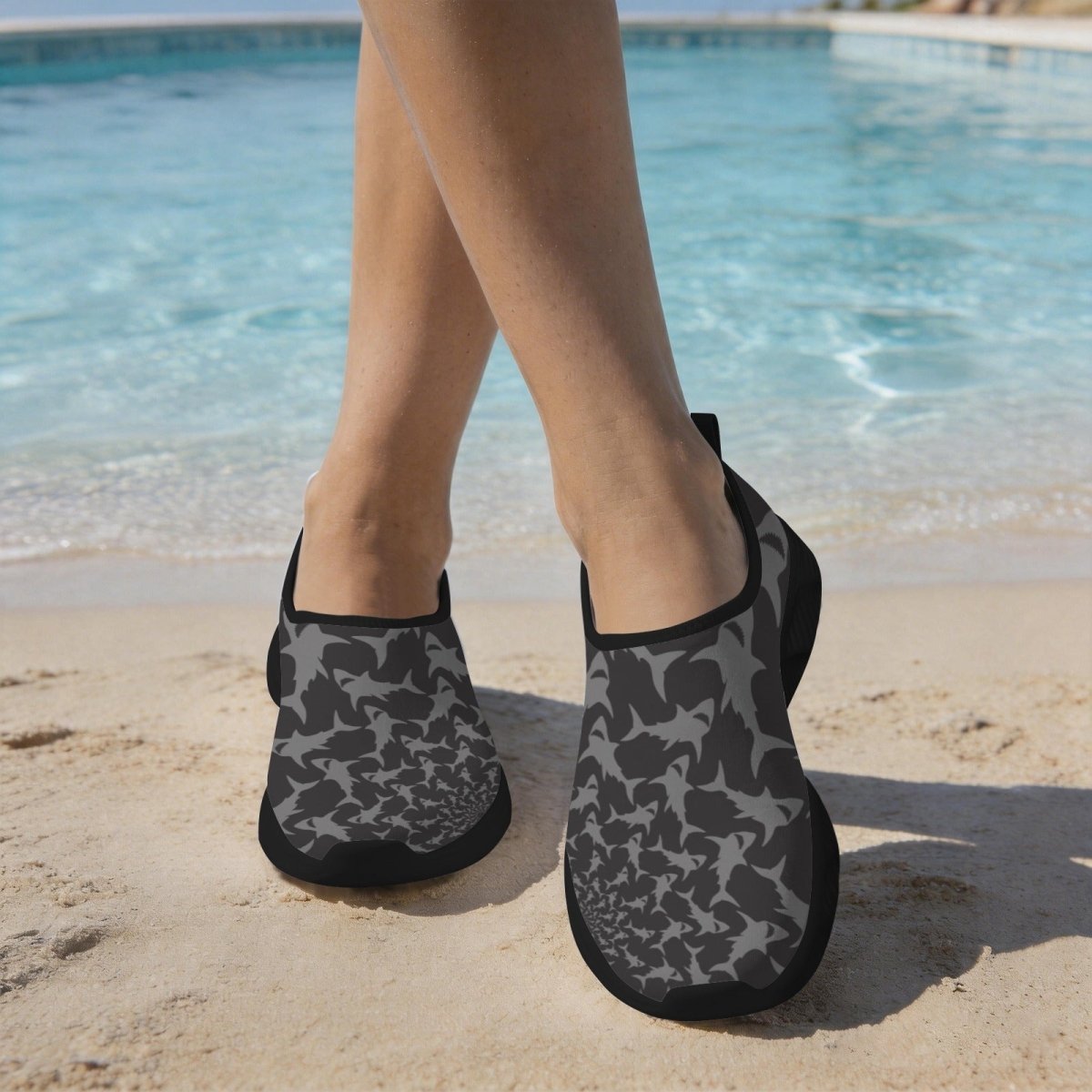 Gray and black shark pattern slip-on water shoes worn by a person standing on a sandy beach with a swimming pool in the background.