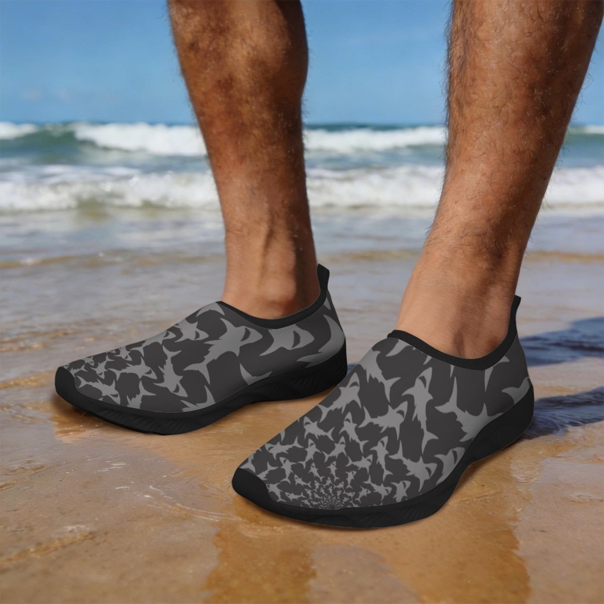 Gray and black Shark Water Slippers worn by a person standing on a wet sandy beach with the ocean waves in the background.