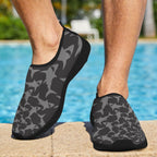 Gray and black shark pattern slip-on water shoes worn by a person standing on a stone pool deck with a swimming pool in the background.