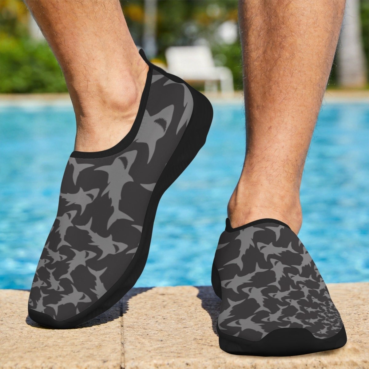 Gray and black shark pattern slip-on water shoes worn by a person standing on a stone pool deck with a swimming pool in the background.