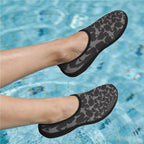 A pair of slip-on water shoes with a dark gray and black shark pattern, shown being worn on feet in a swimming pool.