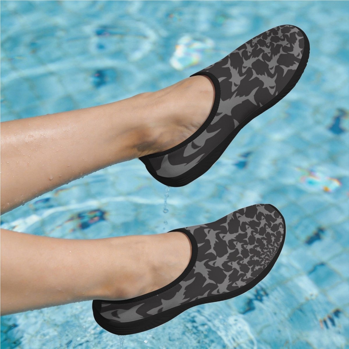 A pair of slip-on water shoes with a dark gray and black shark pattern, shown being worn on feet in a swimming pool.