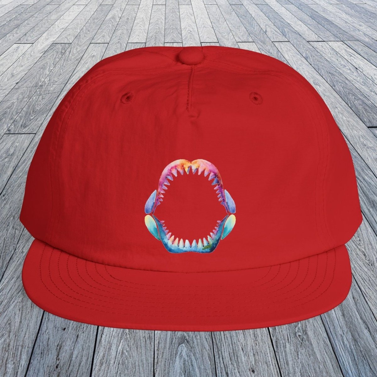 A bright red flat-brim cap with a colorful shark-mouth graphic on the front, displayed on a gray wood plank floor.