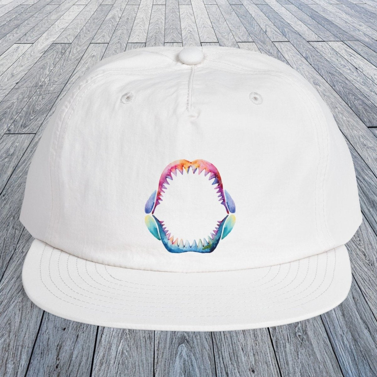A white baseball cap with a colorful shark mouth graphic on the front, displayed on a gray wooden plank surface.