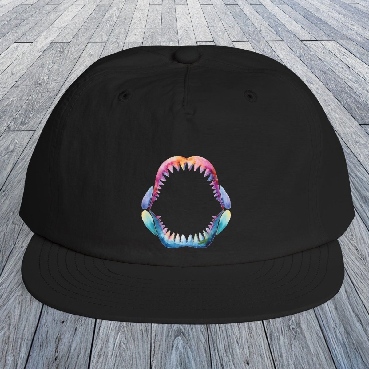 A black flat-brim baseball cap with a colorful shark-mouth graphic on the front, displayed on a gray wood plank surface.