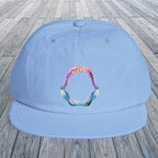 A light blue flat-brim cap with a colorful shark teeth mouth graphic on the front, displayed on a gray wooden plank surface.