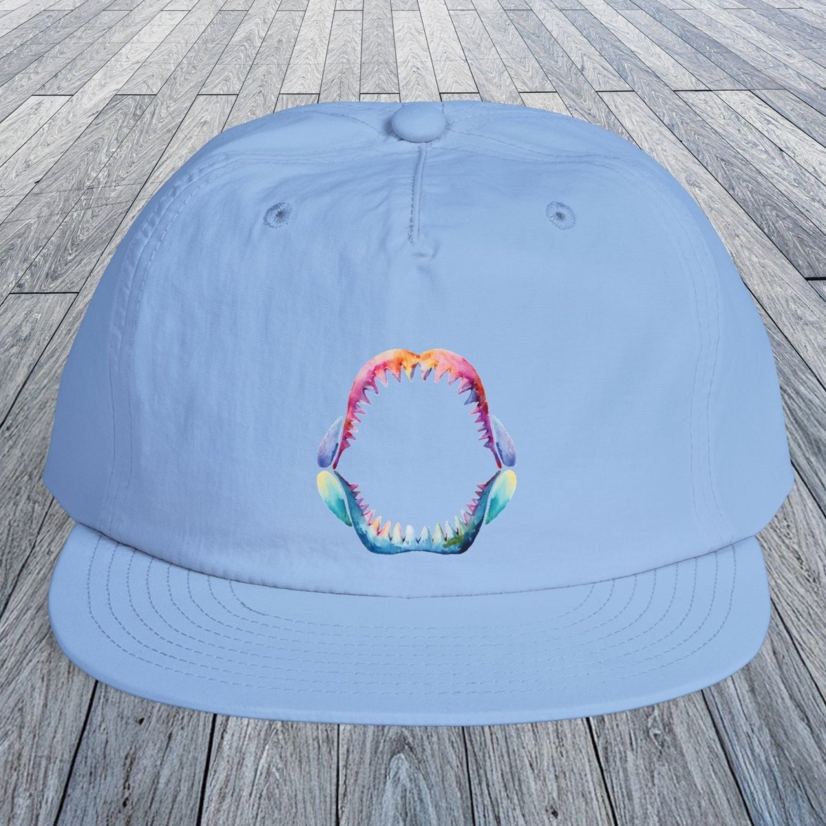 A light blue flat-brim cap with a colorful shark teeth mouth graphic on the front, displayed on a gray wooden plank surface.