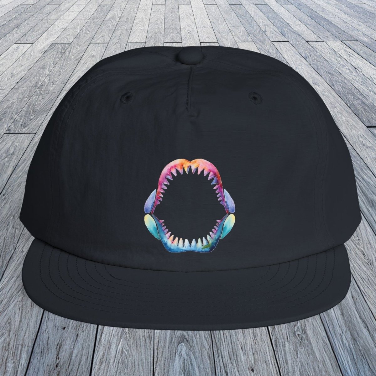 A navy flat-brim surf hat with a colorful shark-mouth graphic on the front, displayed on a gray wooden plank surface.