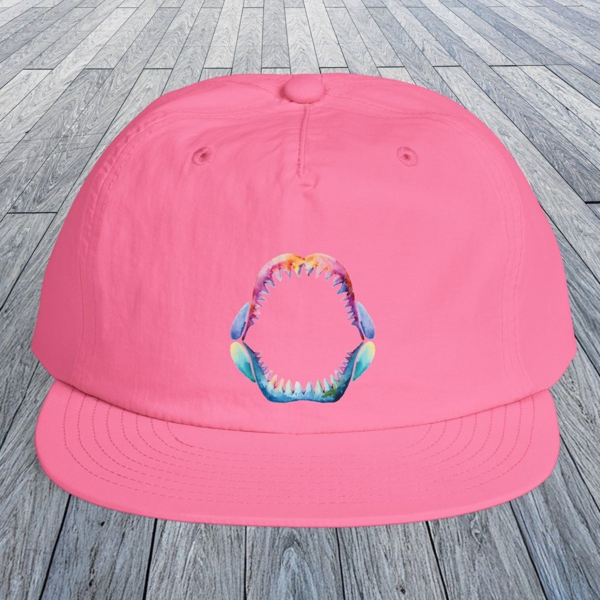 A bright pink flat-brim cap with a colorful graphic of a shark mouth on the front, displayed on a gray wooden plank surface.