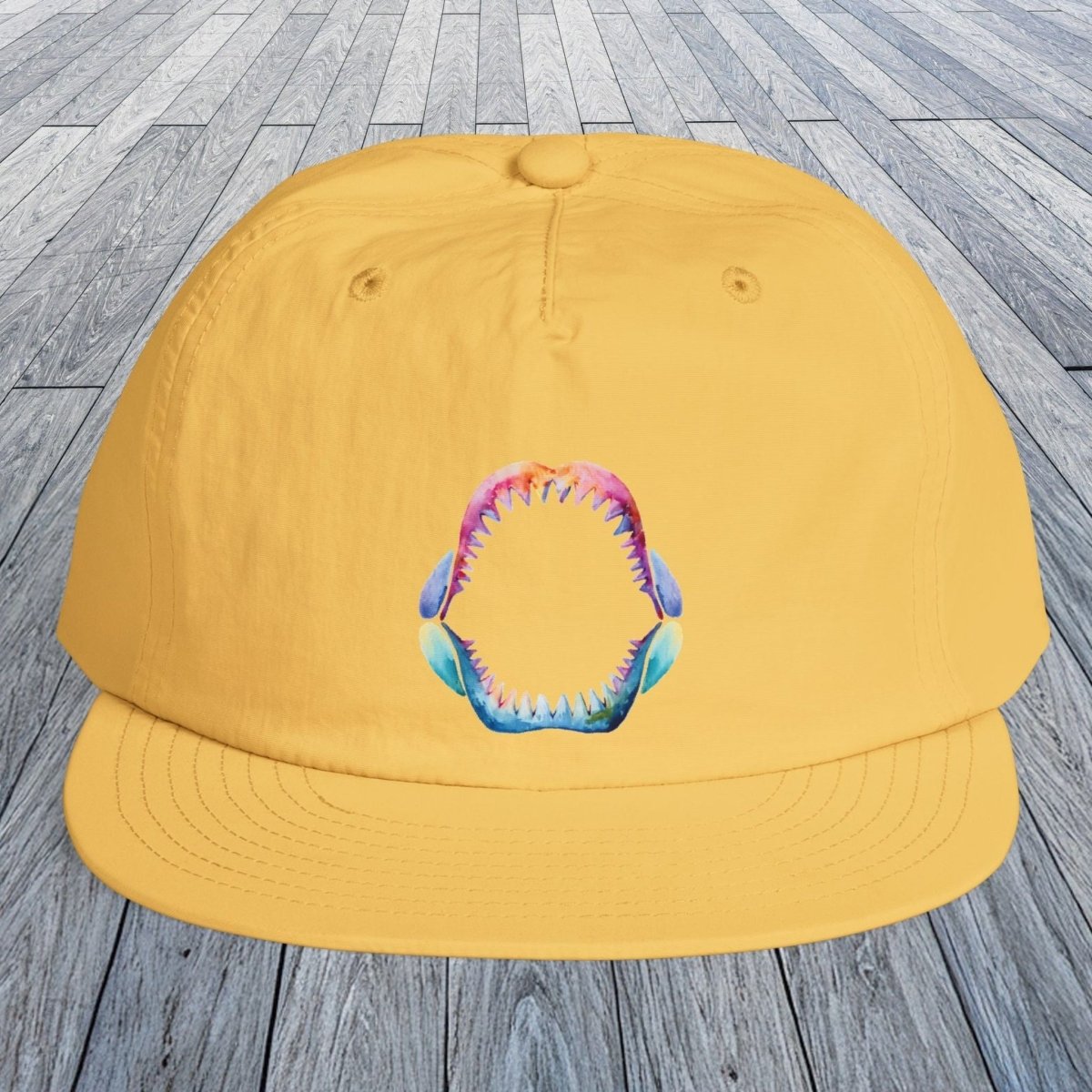 A sunset yellow baseball cap with a colorful shark teeth graphic on the front, displayed on a gray wooden plank surface.