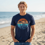 A man standing on a sandy beach wearing a navy blue t-shirt with a retro sunset graphic that reads 'BREATHE IN BREATHE OUT MOVE ON - J.Buffett