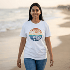 A woman standing on a beach wearing a white graphic t-shirt with a colorful retro circular design that reads 'BREATHE IN BREATHE OUT MOVE ON'.