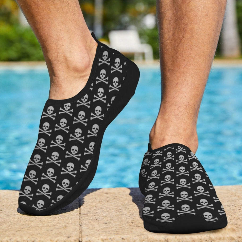 Black slip-on water shoes with a repeating white Skull Bones pattern, shown on a person's feet standing on a poolside deck.