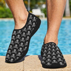 Black slip-on water shoes with a repeating white Skull Bones pattern, shown on a person's feet standing on a poolside deck.