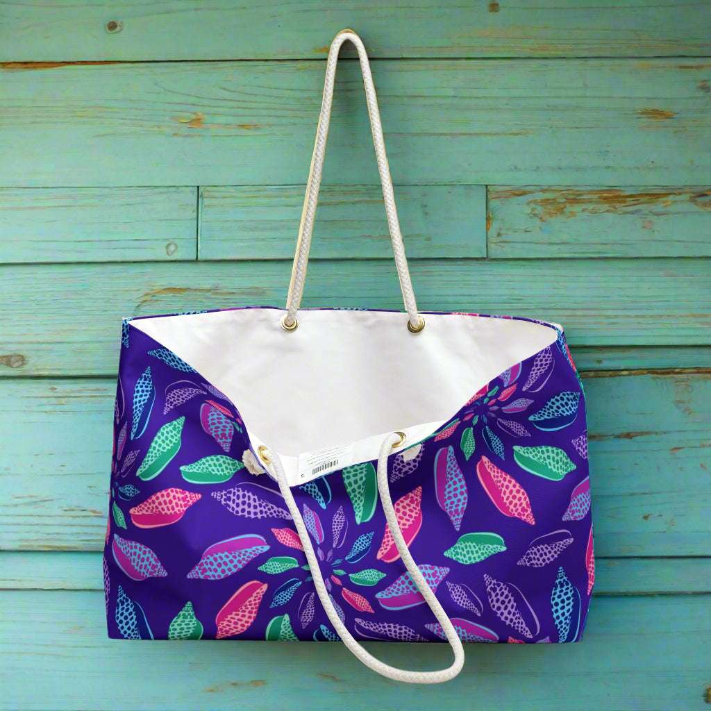 Oversized tote bag with a colorful junonia seashell pattern hanging from a hook in the rustic teal wall of seaside surf shack