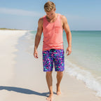 Dude wearing board shorts with colorful junonia seashell pattern and a coral colored tank top walking along the water's edge looking for seashells on Pensacola Beach.