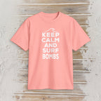 A coral pink short-sleeve t-shirt with a white graphic print reading 'KEEP CALM AND SURF BOMBS' on a distressed light wood background.
